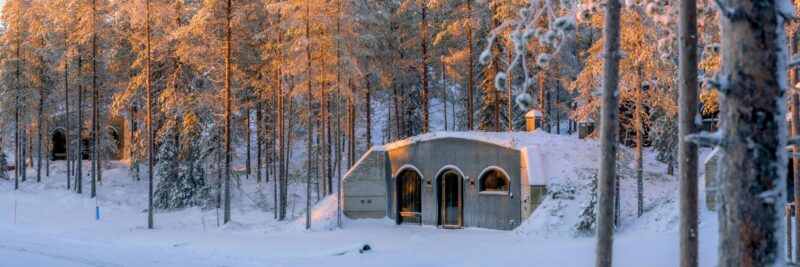 Cabin van de Kaamos Lodge onder een laag sneeuw met zonnestralen die door de bomen schijnen.